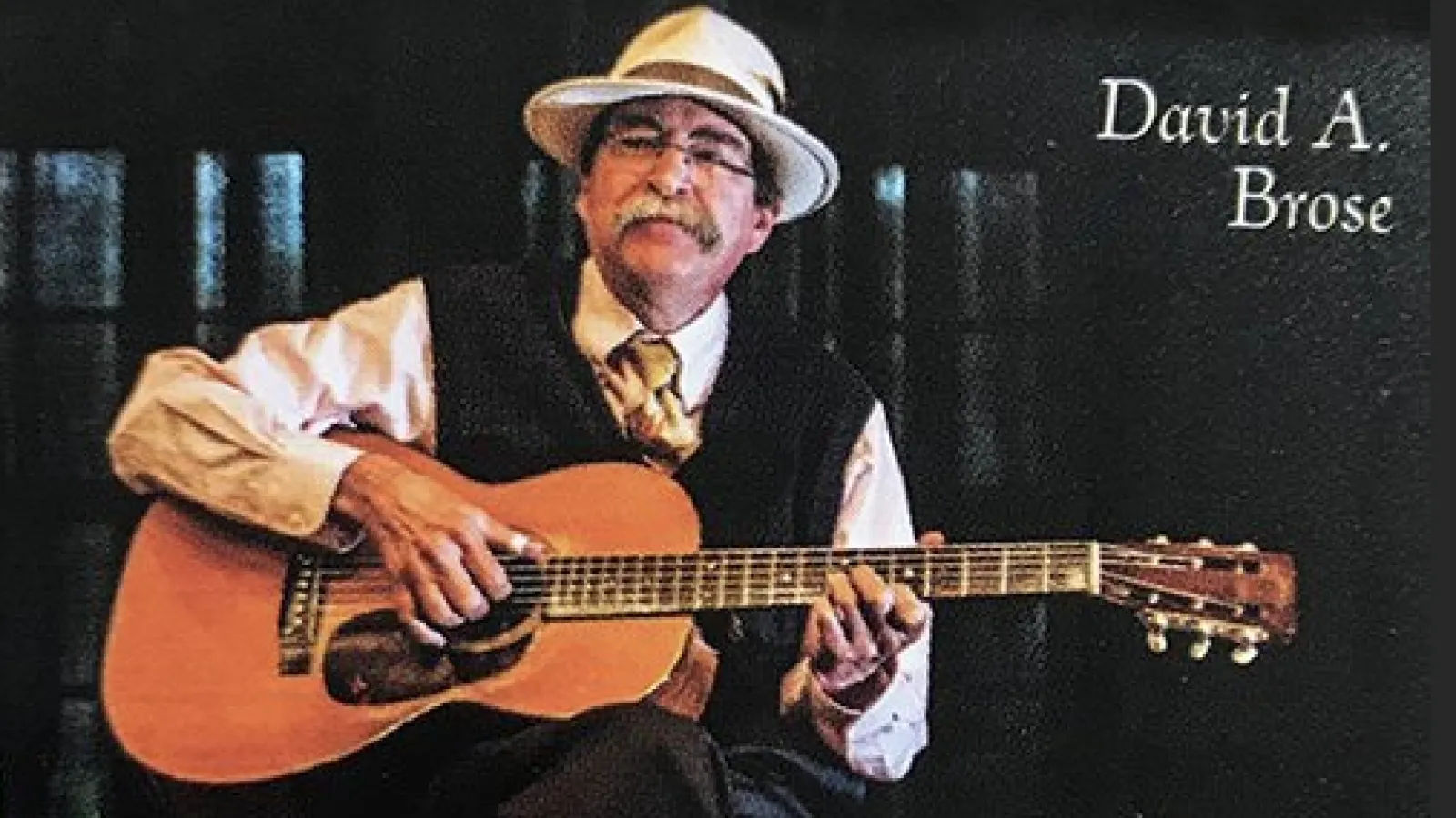 David Brose, a white man with a mustache and glasses wearing a white hat, sitting with a guitar in his lap