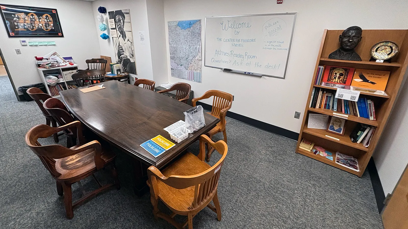 A large wooden table with eight chairs in the center of a room with maps, artwork, and a bookshelf